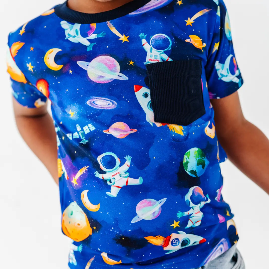 Close-up of a royal blue pocket tee with a navy chest pocket and a space print of astronauts, planets, rockets, and galaxies.